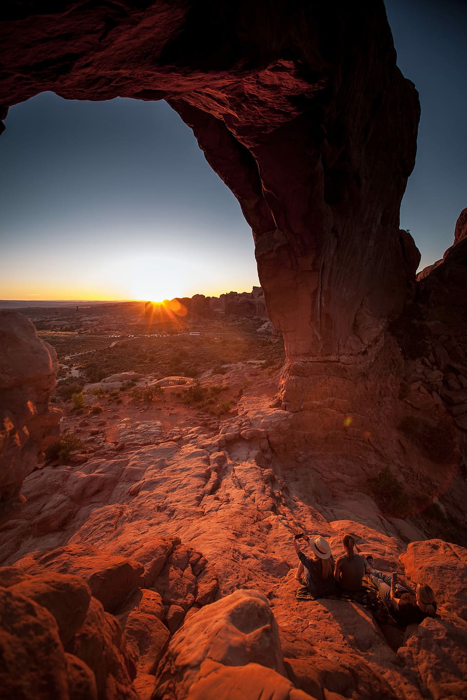 two people sitting on rock below arch facing sun, two person sitting on rock mountain HD wallpaper