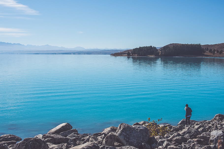 photography of water body, person standing on rocky ground near body of water under blue sky HD wallpaper