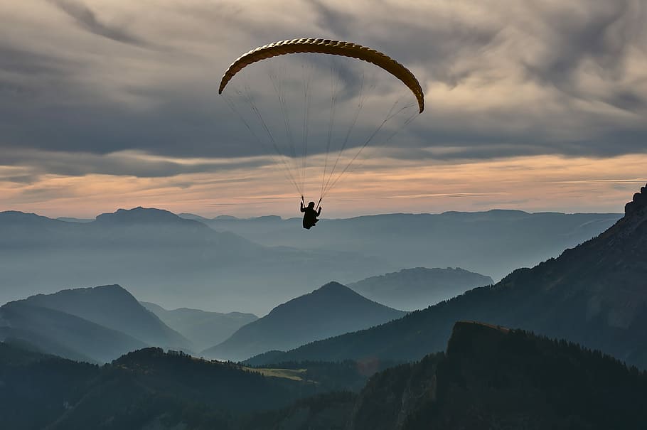 person doing paragliding under cloudy sky, person riding parachute over mountains during daytime