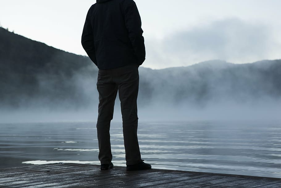 silhouette of man standing beside water across hill during daytime HD wallpaper