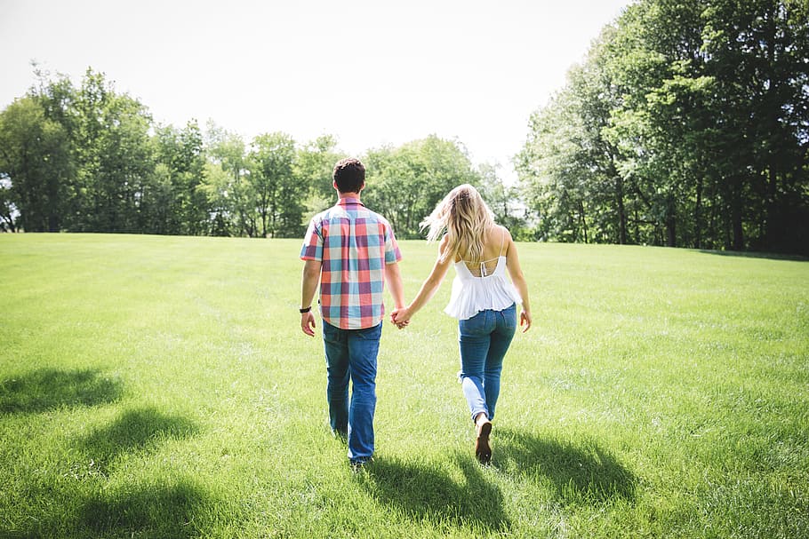 man and woman walking on green grass field surrounded with trees, man and woman holding hands while walking