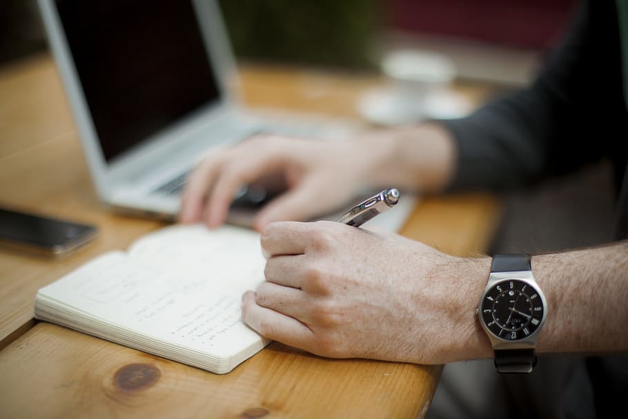 man sitting while writing on notebook, person holding pen writing on table HD wallpaper