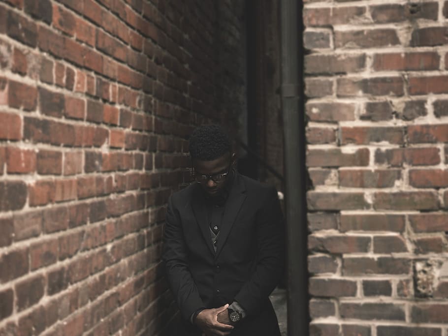 man in black suit standing on corner during daytime, person wearing black suit jacket standing between brown brick walls