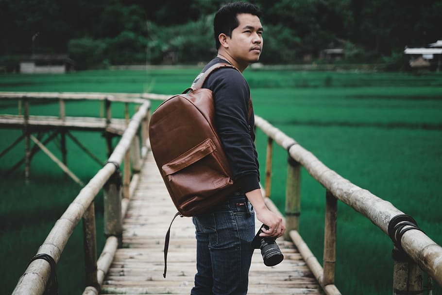 man holding DSLR camera standing on dock, shallow focus photography of man carrying brown leather backpack standing on brown wooden platform during daytime, HD wallpaper