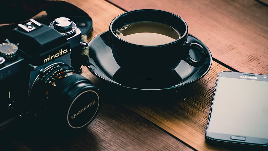 black Minolta camera beside coffee filled black ceramic cup, black ceramic coffee cup on saucer near black Minolta DSLR camera and smartphone