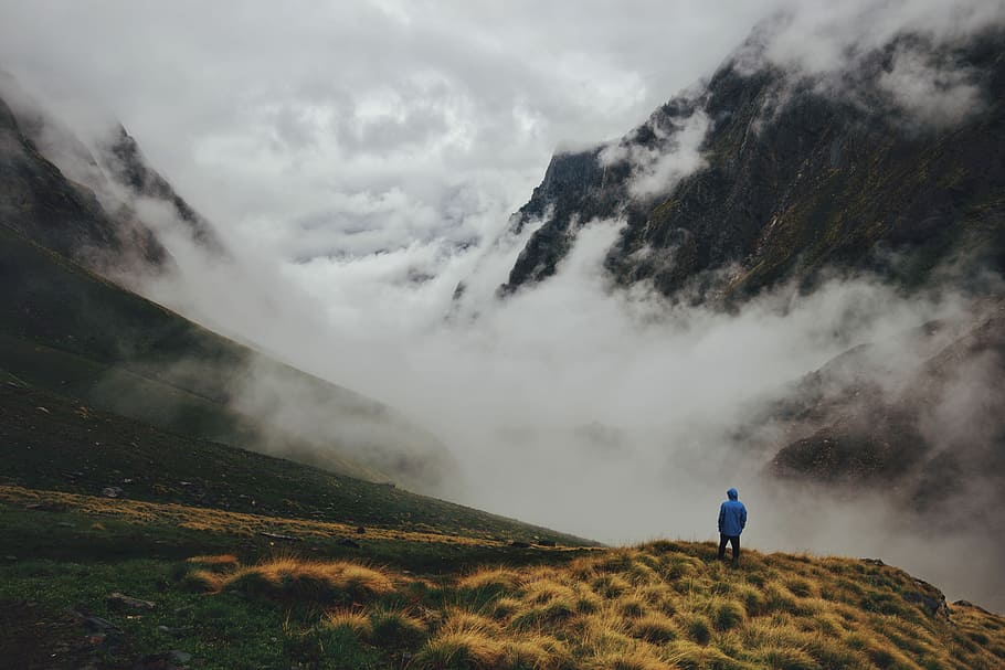 aerial photography of hooded man standing at mountain cliff facing cloudy mountains, man standing on mountain with fogs HD wallpaper
