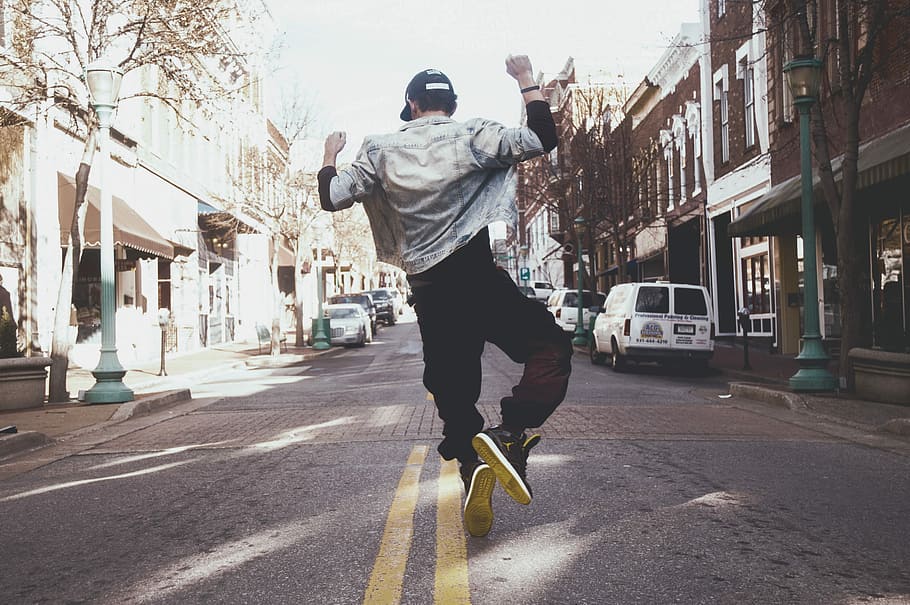 man jumping on the middle of the street during daytime, man wearing white jacket and black pants about to jump on street during daytime
