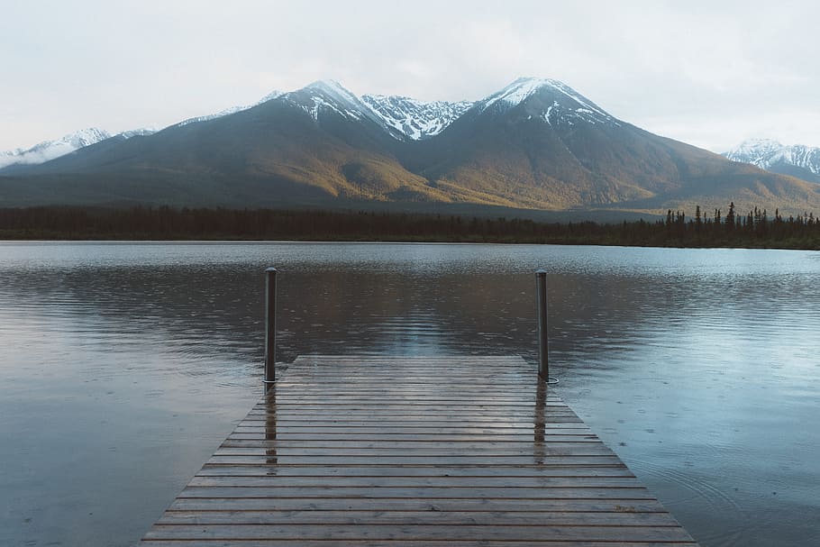 snow-capped mountain at the horizon viewed from lake dock, brown wooden dock on calm body of water in distance brown mountain under white sky at daytimke HD wallpaper