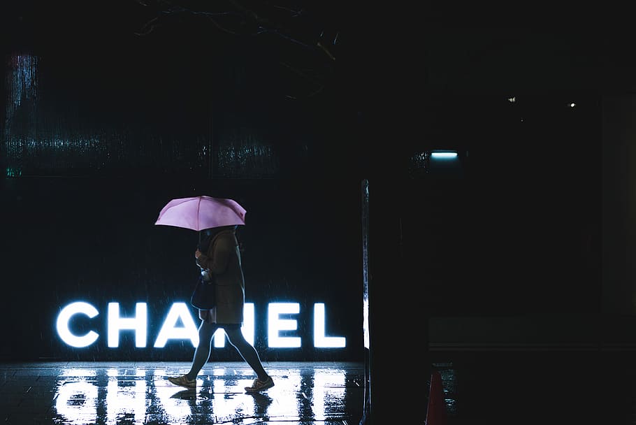 woman walking under umbrella passing by Chanel lighted signage, woman walking beside Chanel LED signage while holding umbrella during nighttime