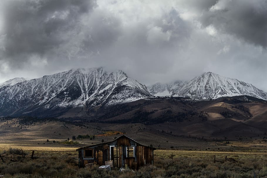 abandoned brown wooden cabin at middle of valley with view of snow-covered mountain range under gray cloud formation during daytime, landscape photography of wooden structure near mountains HD wallpaper