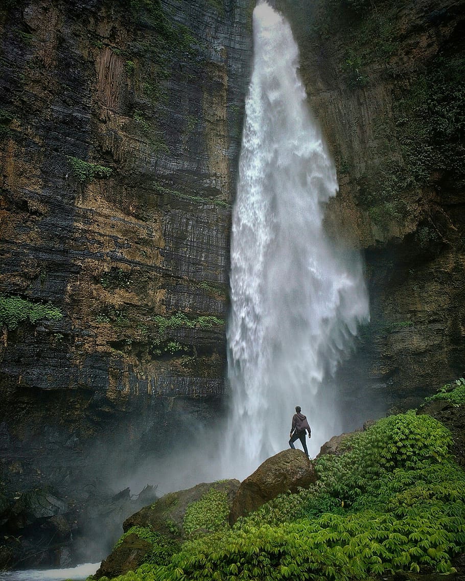 person standing on brown rock formation looking at waterfalls during daytime, person standing in front of waterfalls HD wallpaper