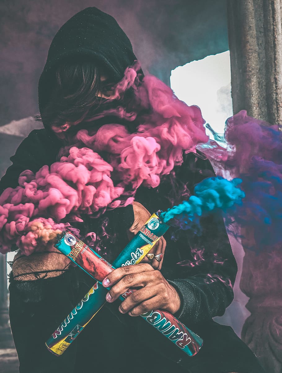 person holding colored smoke, person in black hoodie holding blue and purple rainbow smoke makers