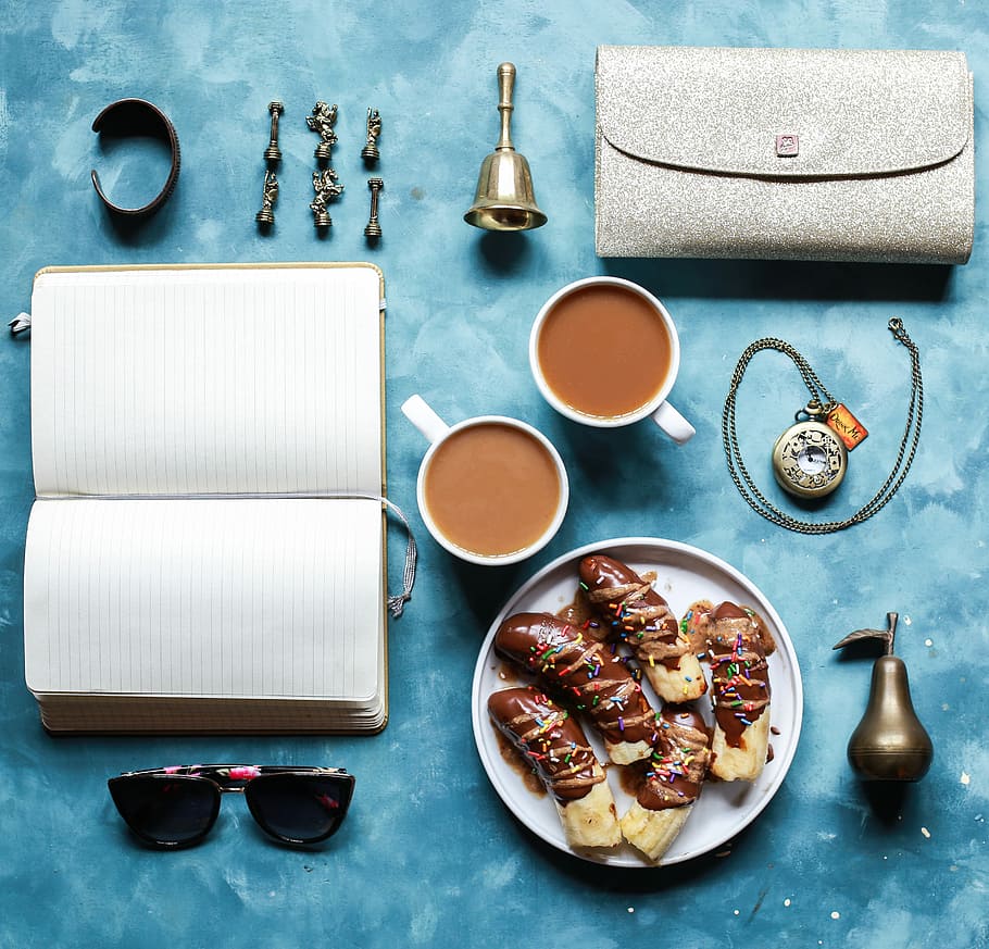 flat lay photography of opened book, coffee mugs, handbag, and sunglasses, coffee mug and plate on blue surface