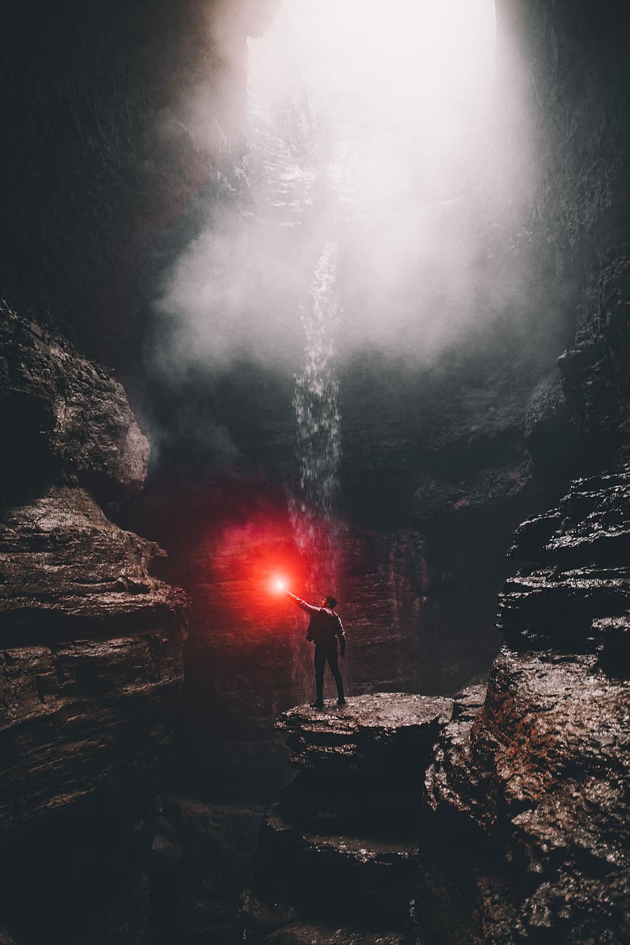man standing on rock formation with water falls, person holding red light inside cave with waterfall