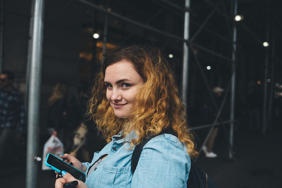 woman holding black smartphone near gray metal frame at daytime, smiling woman wearing blue chambray button-up collared shirt holding blue smartphone near gray scaffolding tunnel HD wallpaper