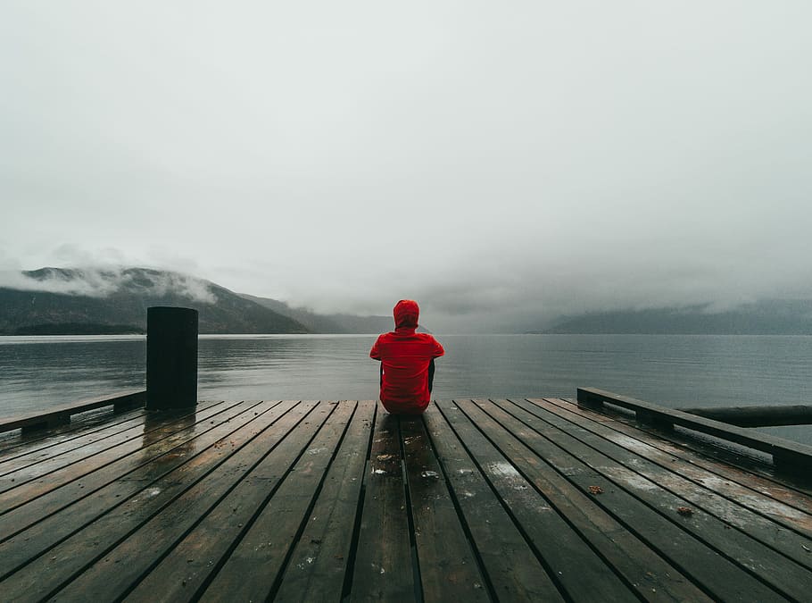 person wearing red hooded jacket sitting on brown and black wooden sea dock near mountains under gray sky during daytime, man sitting in dock looking in the body of water, HD wallpaper