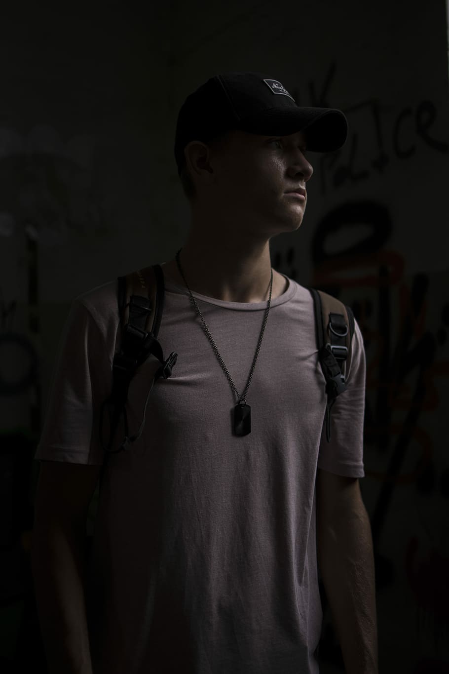 man wearing white T-shirt and backpack standing in a dark lit room, man wearing backpack and cap inside dimmed room
