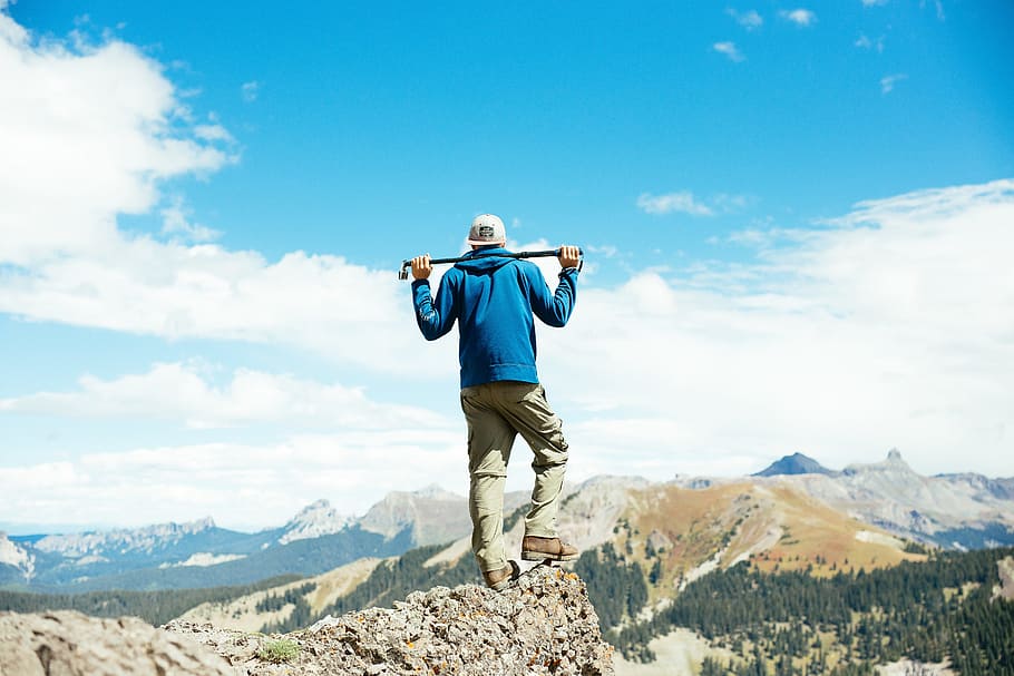 man holding bar on mountain peak during daytime, man standing on stone facing the mountains during day HD wallpaper