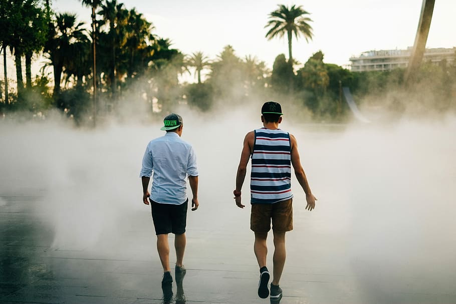 street photography of two men walking in front of water fountain, two men walking on pathway during fog HD wallpaper