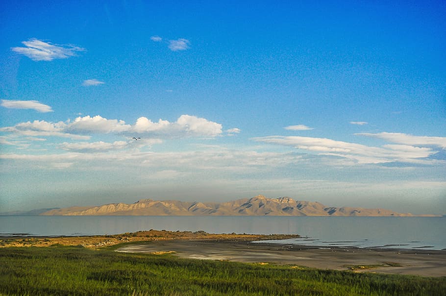 landscape photography of brown hills across calm body of water with distance at green trees under blue cloudy sky HD wallpaper