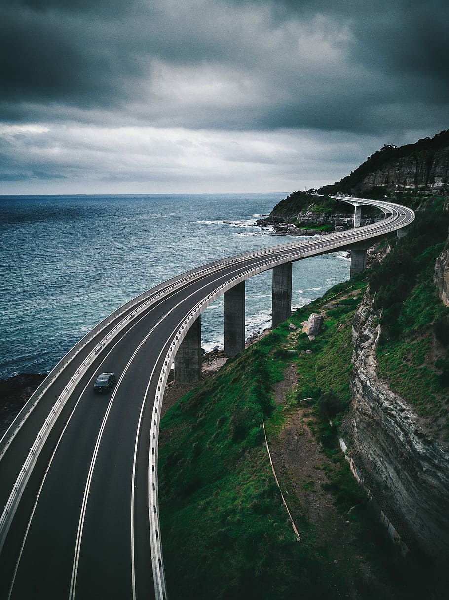 gray concrete road bridge near hill and ocean under white and blue sky, car running on black concrete road HD wallpaper