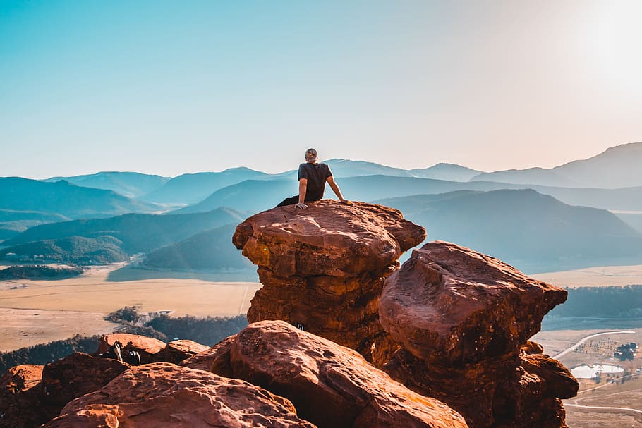 man sitting on rock formation, man wearing black t-shirt on boulders at daytime HD wallpaper