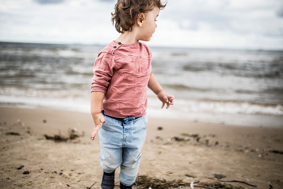 toddler boy standing on seashore, boy wearing pink sweatshirt and gray denim jeans on sand near ocean HD wallpaper