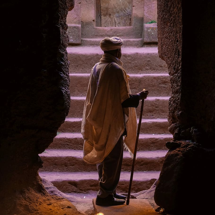 man standing on brown stone cave entrance while holding cane, man wearing beige tuban standing between cave HD wallpaper