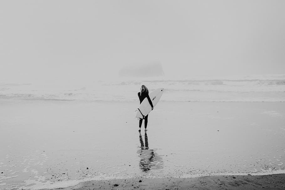 grayscale photo of woman walking on seashore while carrying surfboard, woman carrying surfboard while watching wave of beach rushing to the beach grayscale photography HD wallpaper