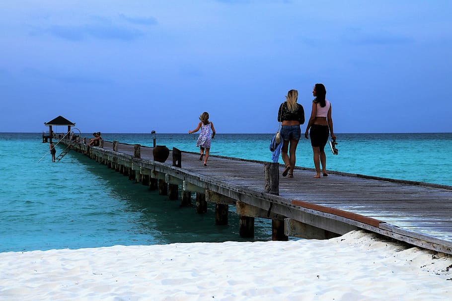 women and child walking on sea dock under blue skies, maldives