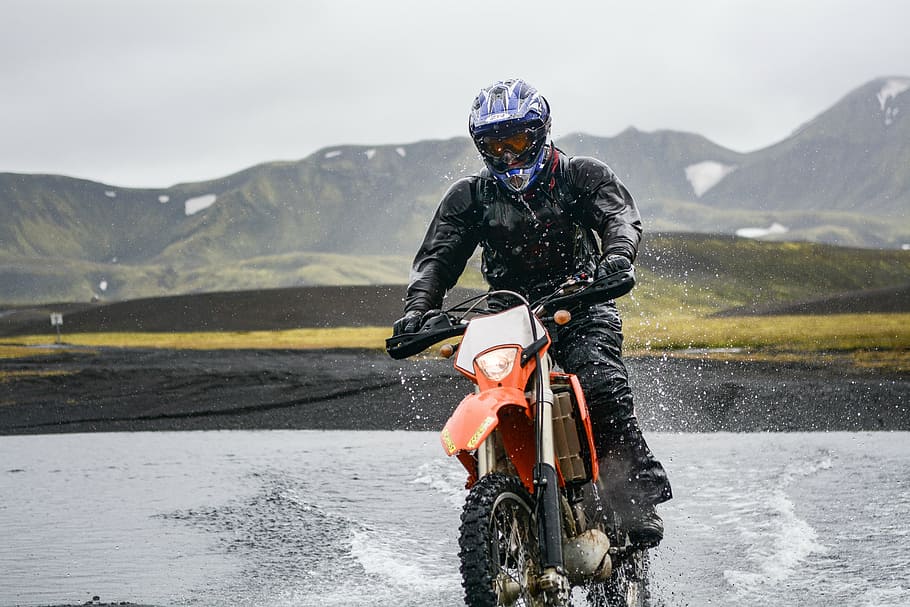 man riding orange dirt bike passing body of water during daytime HD wallpaper