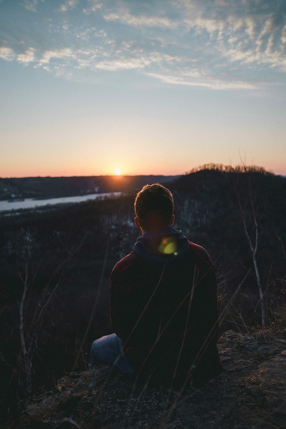 man sitting on grass facing forward on mountain, man in red jacket and blue denim bottoms sitting beside grass watching hill during golden hour HD wallpaper