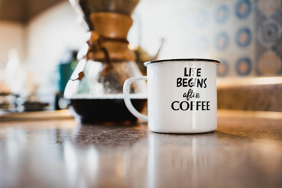 white and black life begins after coffee printed enamel cup beside 1/4 black liquid-filled glass flask on brown wooden panel, white metal mug on table near coffee urn