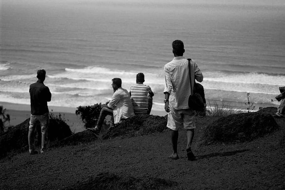 grayscale photo of person walking and facing the seashore, grayscale photo of people on hill near body of water HD wallpaper