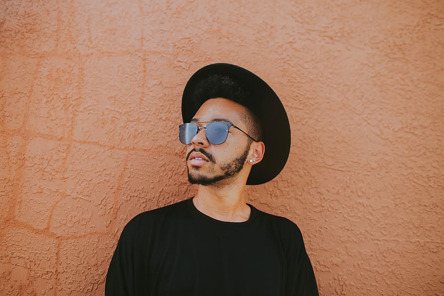 man leaning on brown wall, man in black crew-neck shirt wearing black aviator sunglasses and hat HD wallpaper