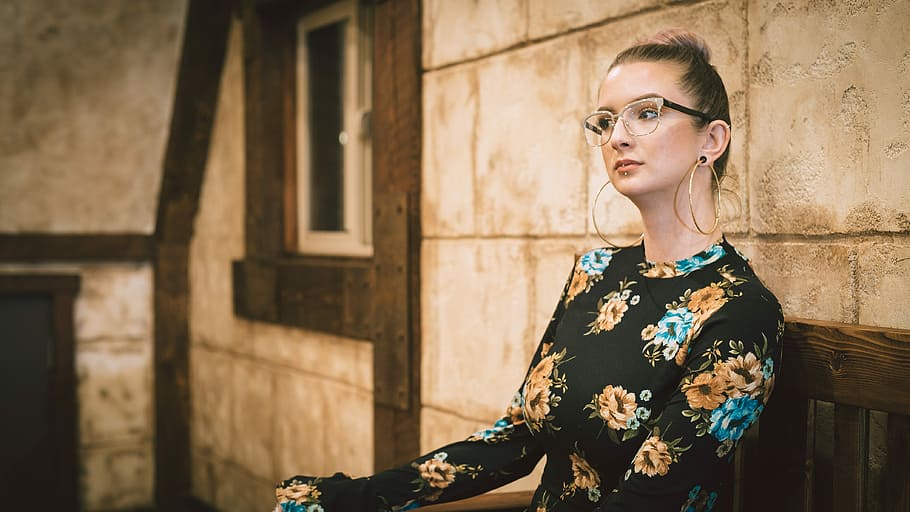 woman in black and multicolored floral long-sleeved top sitting on brown wooden chair beside beige wall, woman sitting on brown wooden bench, HD wallpaper