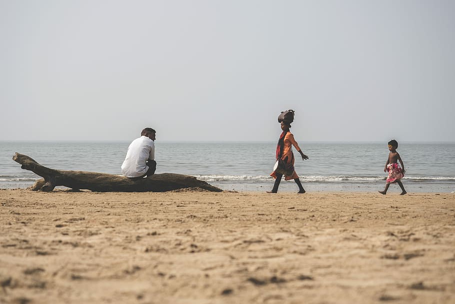 man sitting on drift wood, man sitting on drifwood near woman and young girl walking towards him near ocean water during daytime