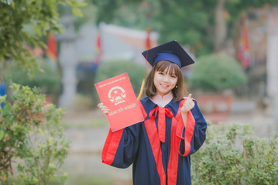 woman wearing black and red academic dress and mortar board holding red book cover near green grass, woman holding red certificate