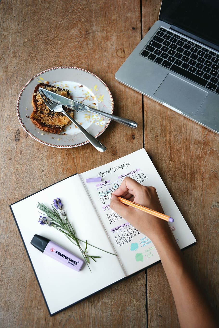 person holding orange pen on to of journal beside Stabilo Boss highlighter, person encircling July calendar on white book beside MacBook Pro and bread on white plate, HD wallpaper