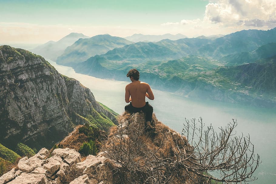 man seating on top of cliff in front of mountains, man on mountain cliff near body of water