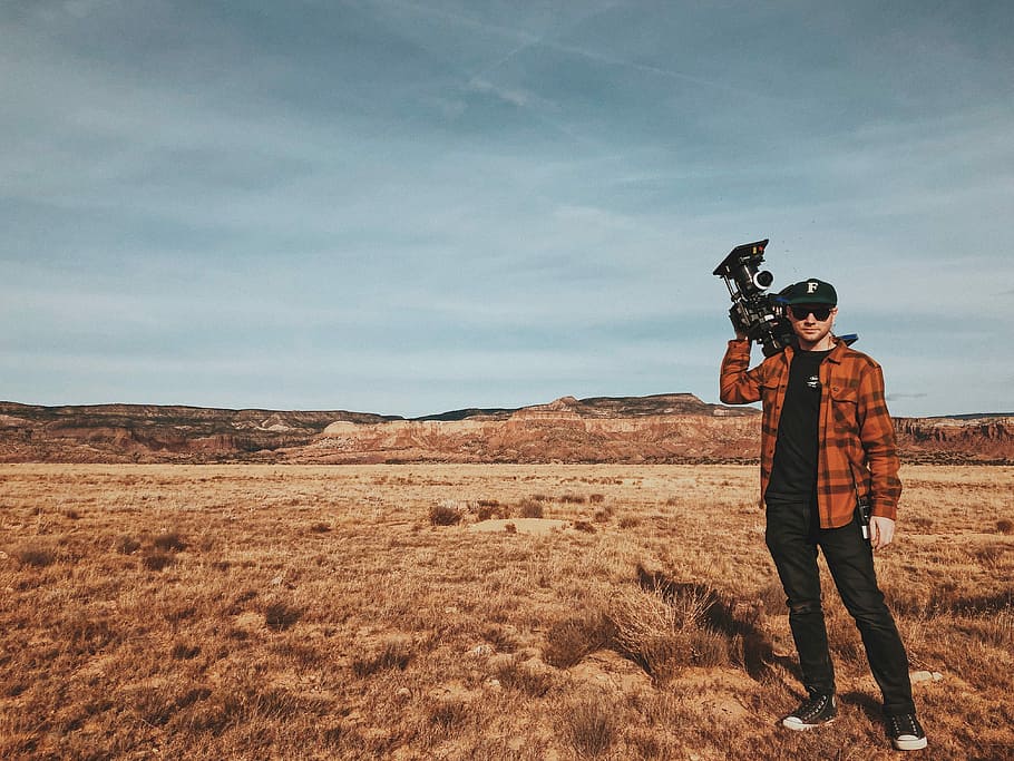 man standing on brown field carrying black recording camera during daytime, man standing on dried grass field while holding camera, HD wallpaper