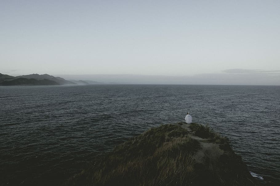 person sitting on a cliff beside a body of water, person sitting on tip of hill during daytime HD wallpaper