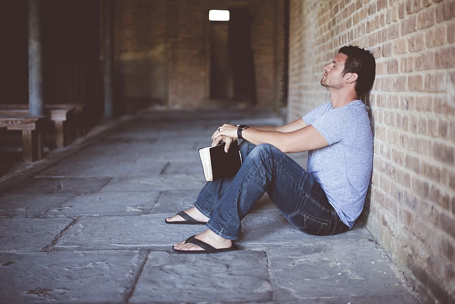 man wearing gray shirt holding book sitting beside brick wall HD wallpaper