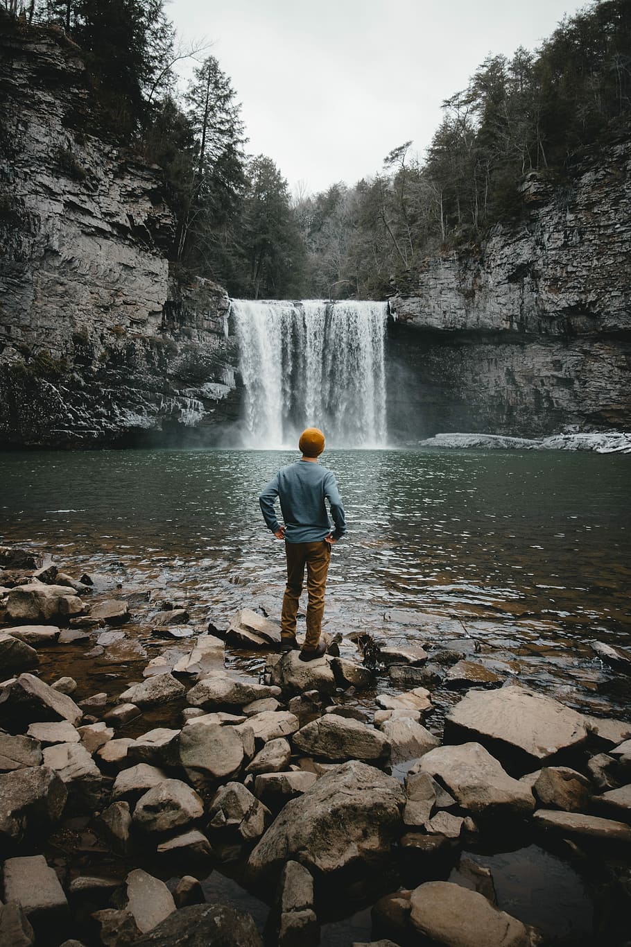 man standing on brown stone facing waterfalls, man facing backward looking at falls HD wallpaper