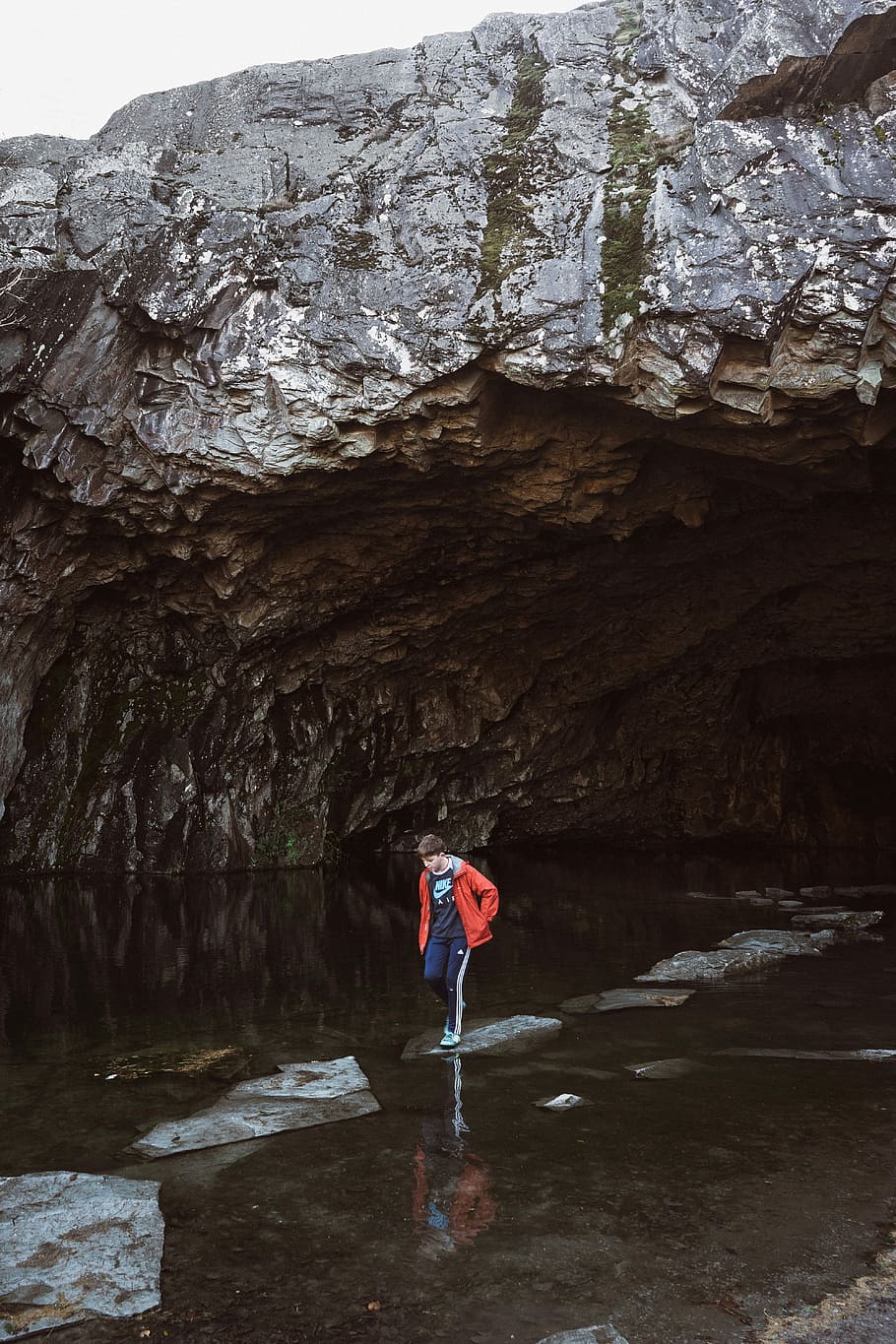 man stepping on stone near cave, boy standing on rock surrounded by body of water near under white sky during daytime HD wallpaper
