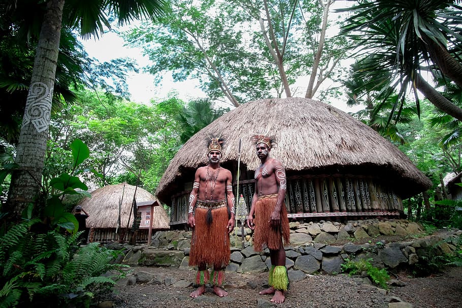 two men standing near house, two man near two brown nipa huts surrounded by trees at daytime