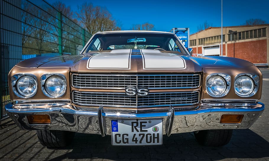 brown and white Chevrolet Chevelle with RE CG 470H license plate parked beside a green fence at daytime HD wallpaper