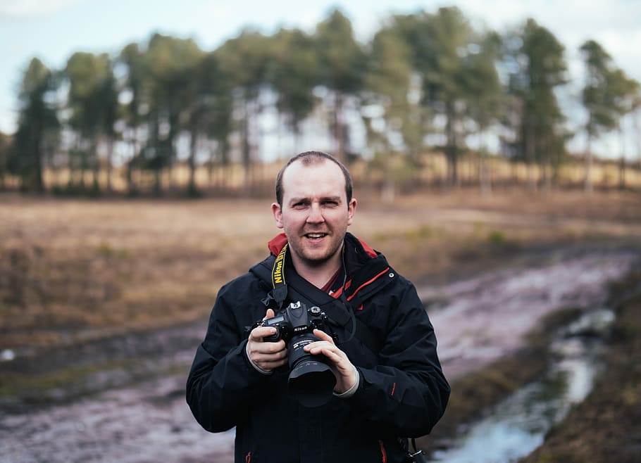 man holding black Nikon DLSR camera while standing on road during daytime, selective focus photography of standing man holding DSLR camera with lanyard HD wallpaper
