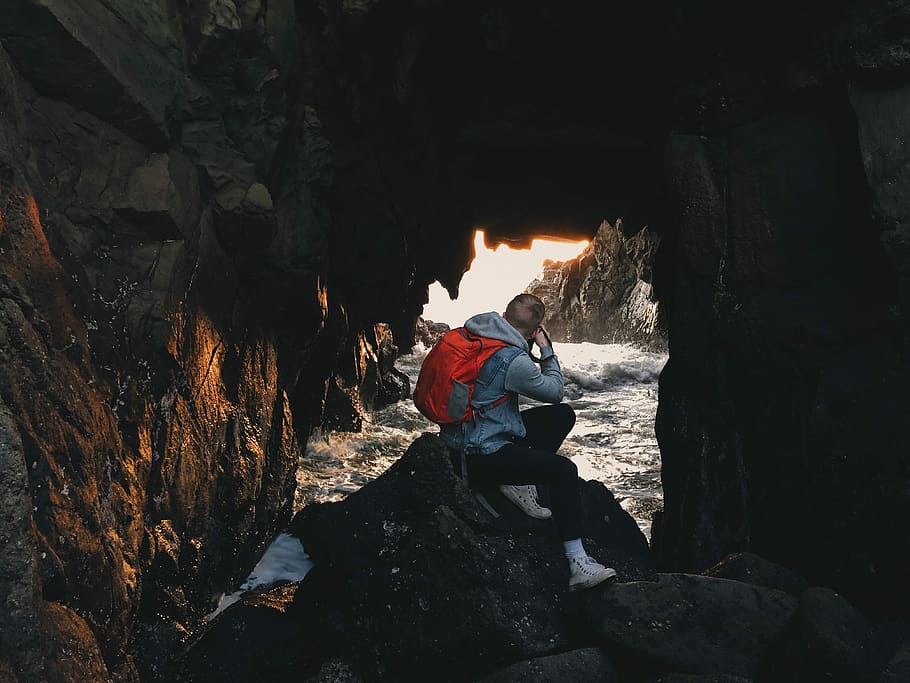 man sitting on gray rock formation, cave, sea, sunlight, sunset HD wallpaper