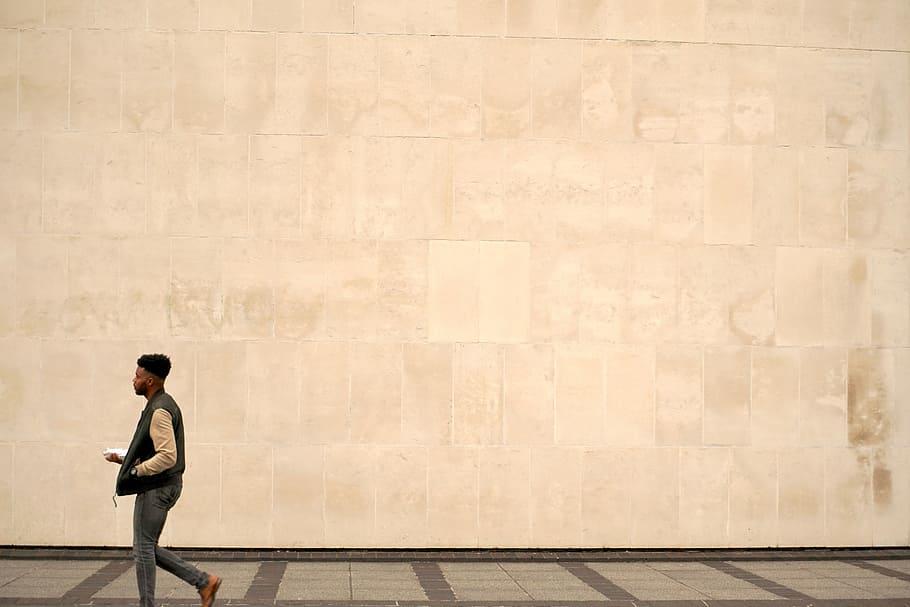 man walking on grey concrete pavement, man holding white device walking near brown concrete wall